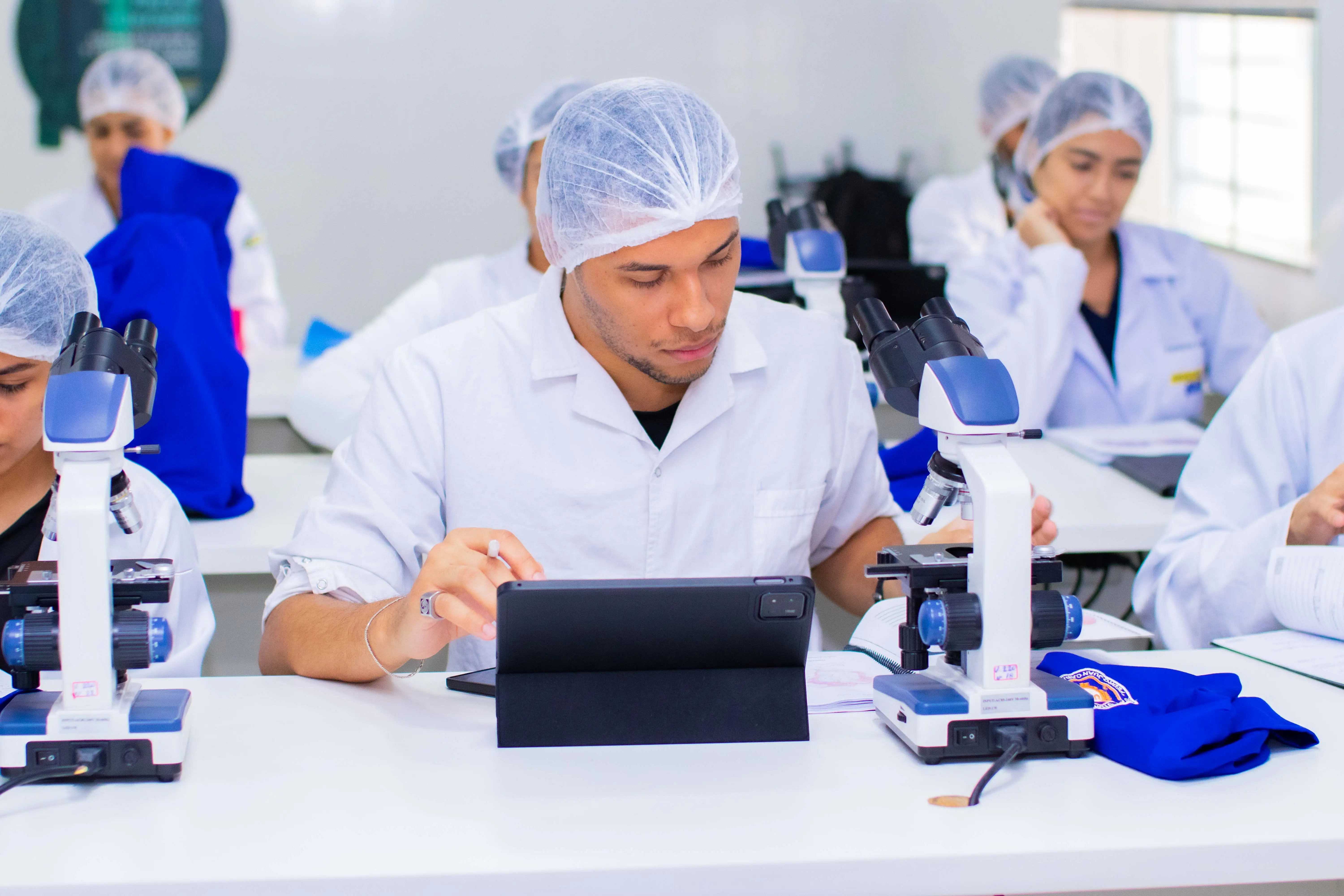 Estudantes de jaleco e touca em bancada de microscópios, em aula de laboratório da UASS-PJC